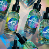 Several clear dropper bottles labeled 'JINX BREAKER' arranged on a reflective iridescent surface. The bottles contain a translucent blue-green liquid and have black dropper caps with pink-to-purple gradient labels.