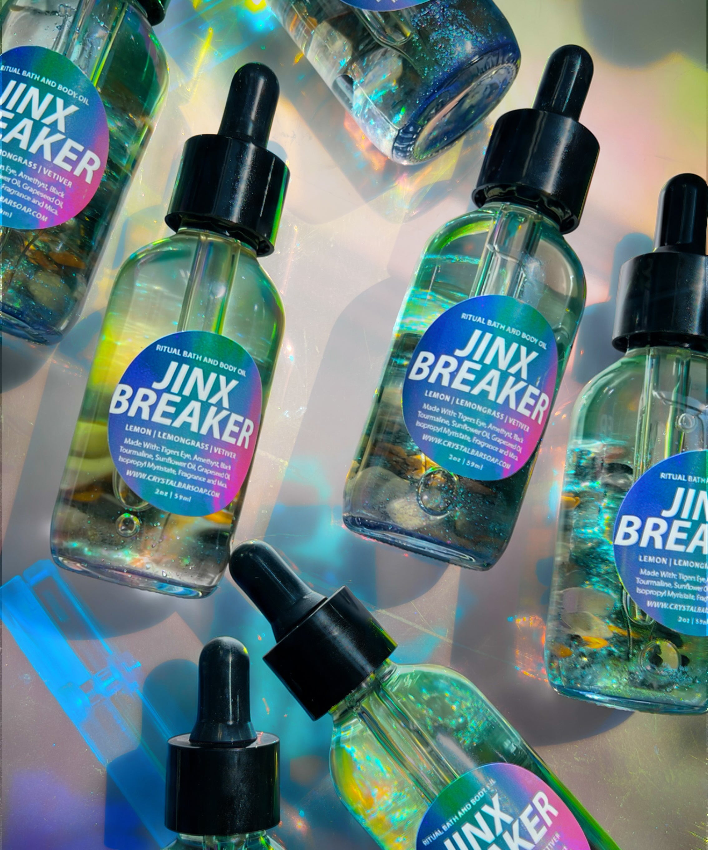Several clear dropper bottles labeled 'JINX BREAKER' arranged on a reflective iridescent surface. The bottles contain a translucent blue-green liquid and have black dropper caps with pink-to-purple gradient labels.