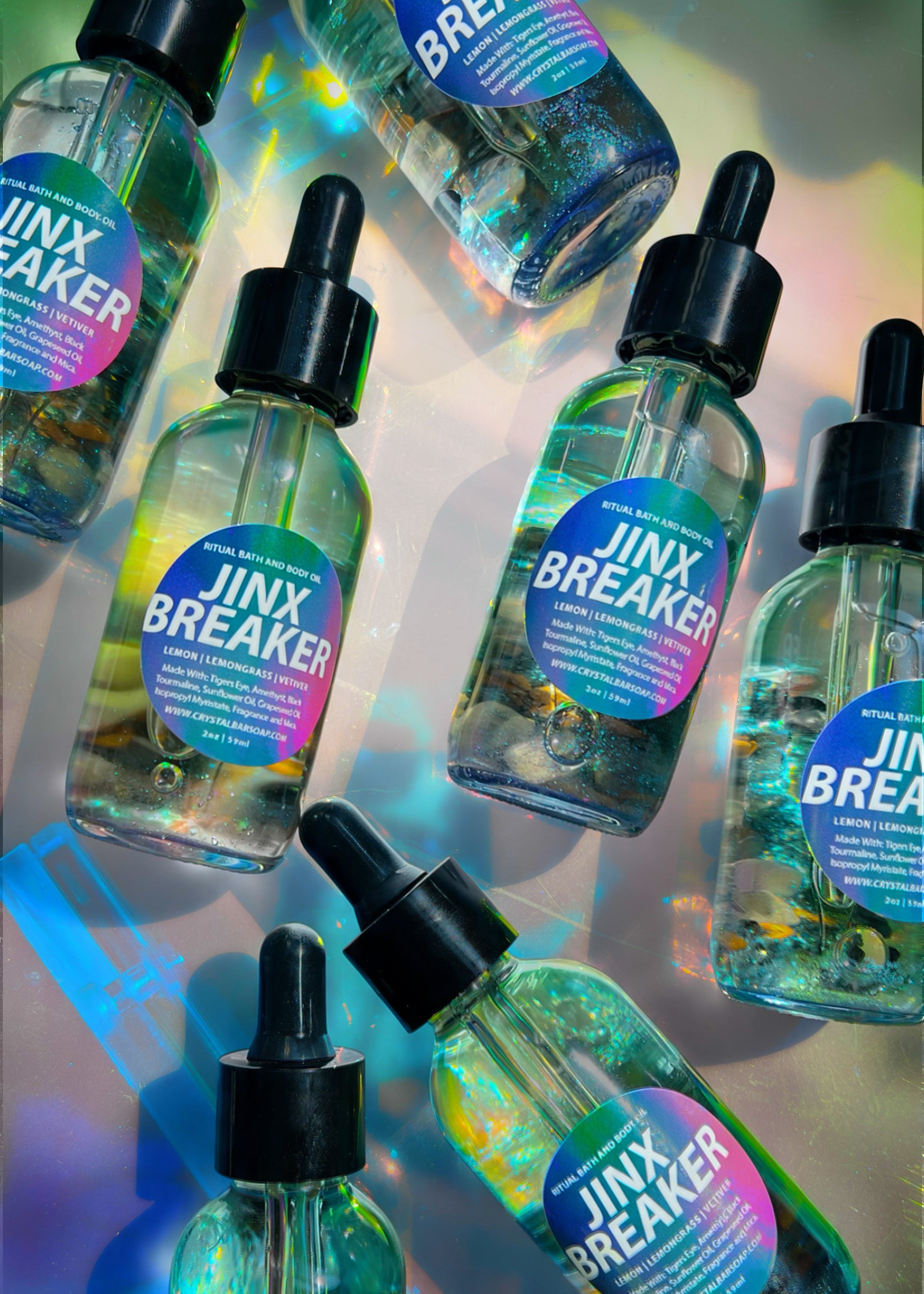 Several clear dropper bottles labeled 'JINX BREAKER' arranged on a reflective iridescent surface. The bottles contain a translucent blue-green liquid and have black dropper caps with pink-to-purple gradient labels.